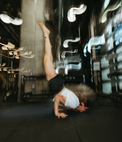 A person balances on their arms in a yoga pose, legs extended upward. The background is blurred with swirling lights, suggesting motion and focus in an Austin climbing gym or fitness studio setting.