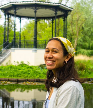 A woman with long dark hair, wearing a yellow-and-white headscarf and hoop earrings, smiles while standing by a pond with greenery and a gazebo in the background.