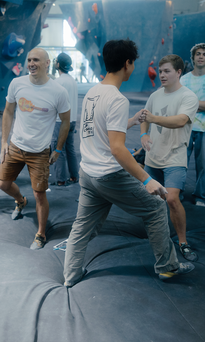 A group of young men in casual clothes stand and interact on the padded floor of the Austin Bouldering Project, with climbing walls and holds visible in the background—perfect for Group Climbing Reservations.