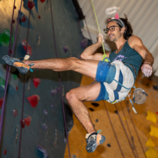 A man wearing a tank top, shorts, glasses, and a headband with fake angel wings smiles while climbing an indoor rock wall. He is suspended by a rope, with one leg raised and chalk on his hand.