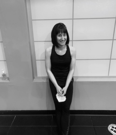A woman with short dark hair, wearing a sleeveless black top and black pants, stands and smiles while leaning against a tiled wall indoors, holding a white cloth in her hands. The photo is in black and white.