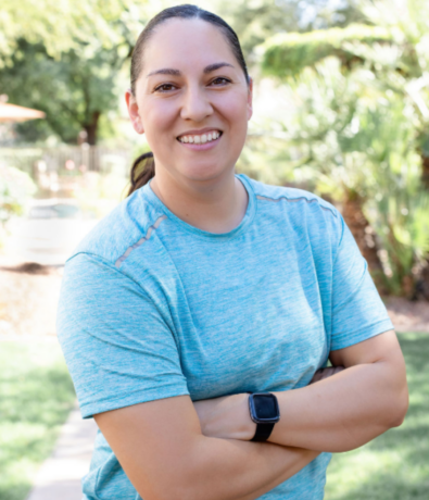 A person wearing a light blue athletic shirt and a smartwatch stands outdoors with arms crossed, smiling, with greenery and trees blurred in the background.