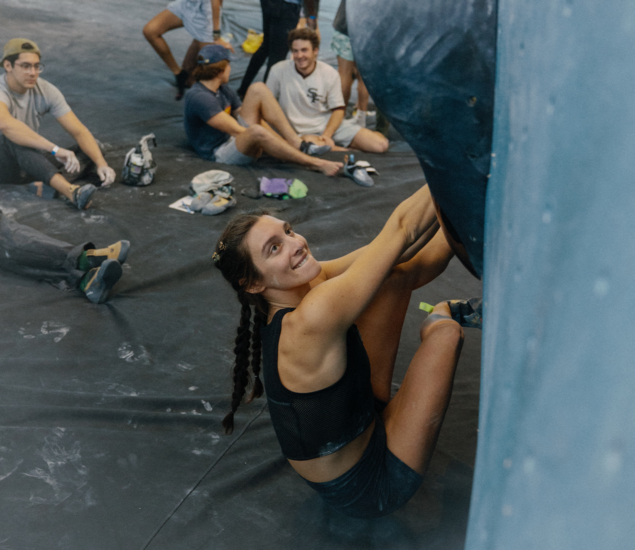 A woman with braided hair in athletic clothing smiles while preparing to climb at Height Fight in Austin, surrounded by other climbers sitting and watching on padded flooring.