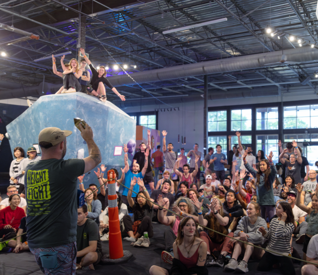 A large crowd of people indoors, many sitting and raising their hands, watch a man in a green shirt at the front; two people sit atop a climbing wall, also raising their arms during the lively Height Fight event in Austin. The atmosphere is energetic.