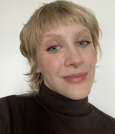 A person with short, blonde hair and blue eyes smiles softly at the camera. They are wearing a dark turtleneck and are posed in front of a plain, light-colored background.
