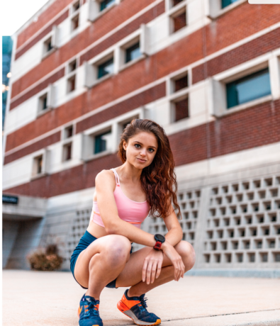 A woman in athletic wear crouches on a Fremont sidewalk outside a brick building with upper walls in view, looking confidently at the camera. She wears a pink crop top, navy shorts, and blue running shoes with orange accents.