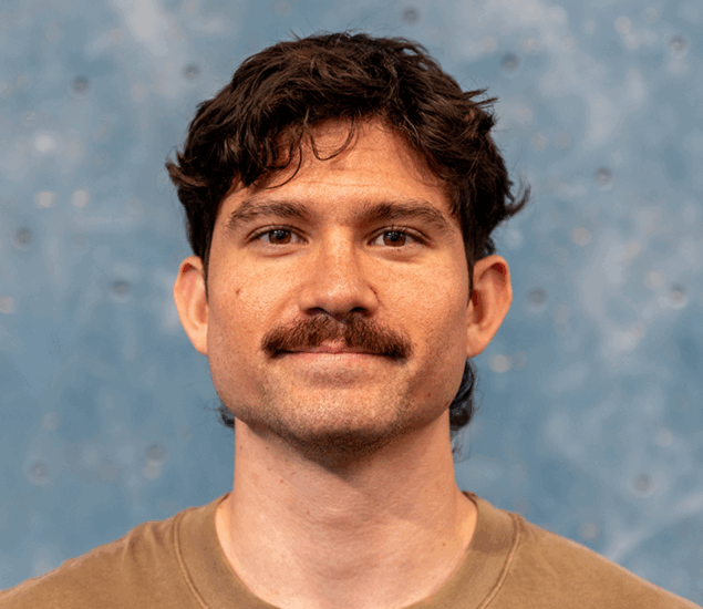 A man with wavy dark hair and a thick mustache is facing forward and smiling slightly. He is wearing a light brown T-shirt. The background, a blue speckled wall, hints at his connection to Personal Climbing Coaching Austin.