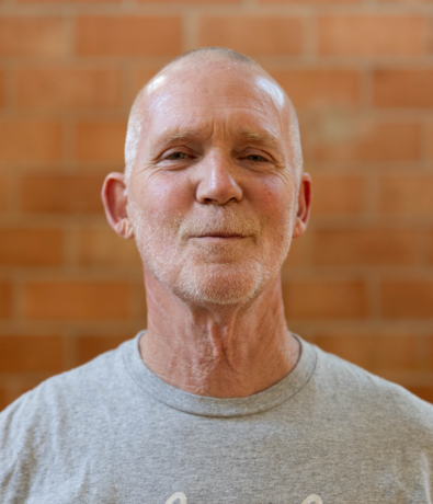 An older man with short, light hair and a trimmed beard wears a gray t-shirt and stands in front of a brick wall at an Austin climbing gym, looking at the camera with a slight smile.