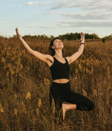 A woman in black workout clothes kneels on one knee in a grassy field, arms raised joyfully, smiling and looking up at the sky under scattered clouds.