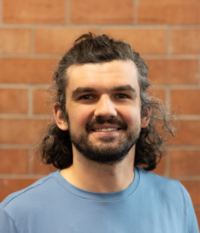 A man with curly brown hair and a beard smiles at the camera. He is wearing a light blue shirt, standing in front of a red brick wall at an Austin climbing gym.