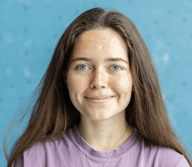A young woman with long brown hair and light eyes, wearing a purple shirt and a necklace, smiles softly at the camera. The background is light blue with small dark dots, evoking the supportive spirit of Personal Climbing Coaching Austin.
