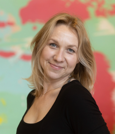A woman with blonde hair and blue eyes smiles at the camera, wearing a black top. The background features a blurry, colorful map with shades of red, pink, green, and white.