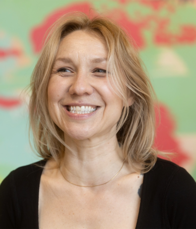 A smiling woman with blonde hair wearing a black top and a thin necklace, standing in front of a colorful, abstract background with pink and green tones.