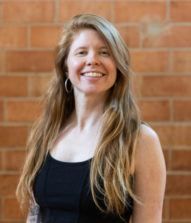 A woman with long, reddish-blonde hair wearing a black sleeveless top smiles at the camera. She stands in front of a wall made of reddish-brown bricks at an Austin Climbing Gym.