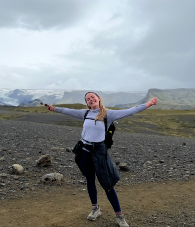 A smiling woman stands outdoors on a rocky trail with her arms outstretched, wearing hiking clothes and a backpack. Mountains and cloudy sky appear in the background.
