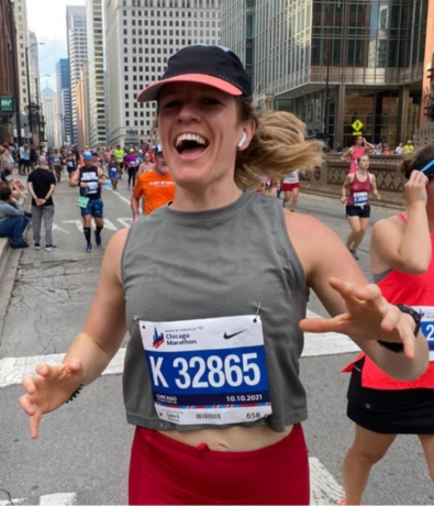 A smiling runner wearing a race bib labeled K 32865 participates in the Chicago Marathon. City buildings and other runners appear in the background on a city street.