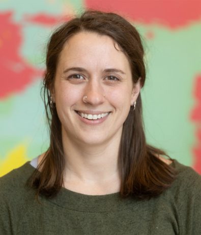 A woman with long brown hair, wearing a green top and small hoop earrings, smiles at the camera. She has a nose ring and is posed in front of a colorful, blurred background.