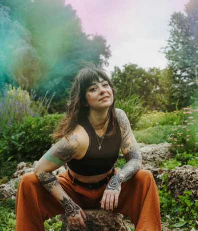 A woman with tattoos on her arms and hands sits on a rock outdoors, surrounded by greenery and wildflowers. She wears orange pants and a black sleeveless top, with glitter on her face and a soft, colorful light effect in the background.