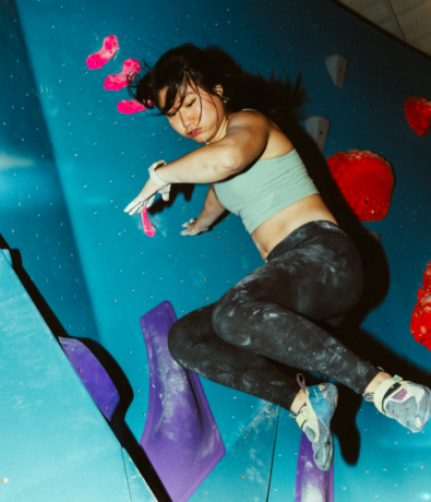 A woman with chalked hands and climbing shoes is mid-movement on an indoor climbing wall at an indoor rock climbing gym, wearing a green sports top and black leggings, with colorful holds beneath and around her.