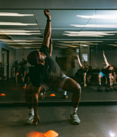 A group of people in workout clothes stretch together in an indoor rock climbing gym, reaching one arm upward while balancing. Their movements are reflected in a large mirror behind them, and orange cones are on the floor.