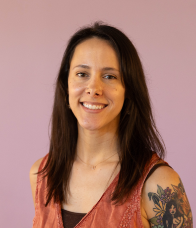 A woman with long brown hair smiles at the camera in a sleeveless, rust-colored top, showing off a colorful tattoo on her upper arm. The light purple background hints at the vibrant style of an Austin Climbing Gym.