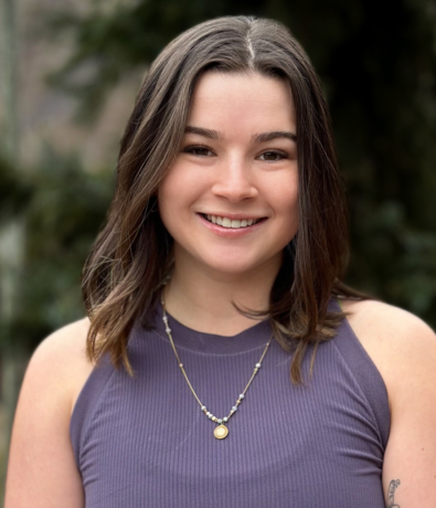 A young woman with shoulder-length brown hair and a sleeveless purple top smiles at the camera. She wears two layered necklaces and stands outdoors with blurred greenery in the background.