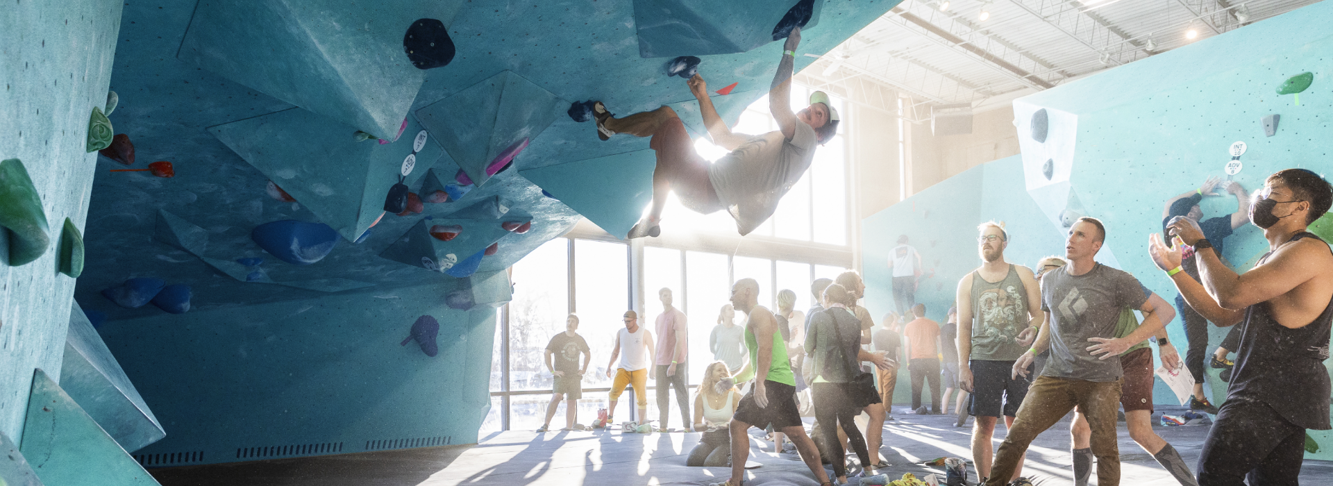 A group of people watch as a climber hangs from a blue indoor bouldering wall in a sunlit climbing gym; some people stand, others sit, with climbing gear scattered around.