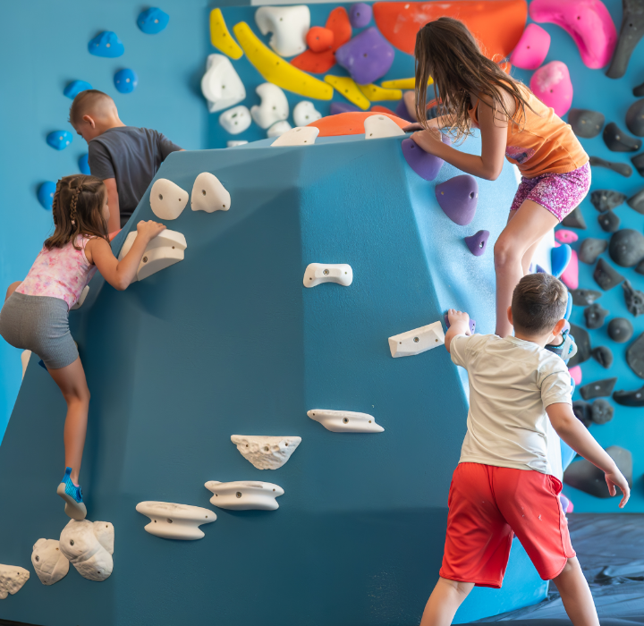 Children climb on blue indoor bouldering walls with colorful climbing holds, while others stand nearby, in a bright play area with natural light coming through large windows.