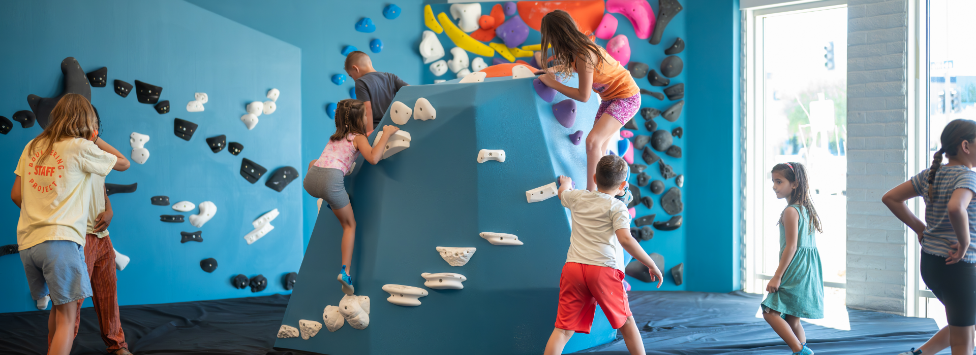 Children climb on blue indoor bouldering walls with colorful climbing holds, while others stand nearby, in a bright play area with natural light coming through large windows.