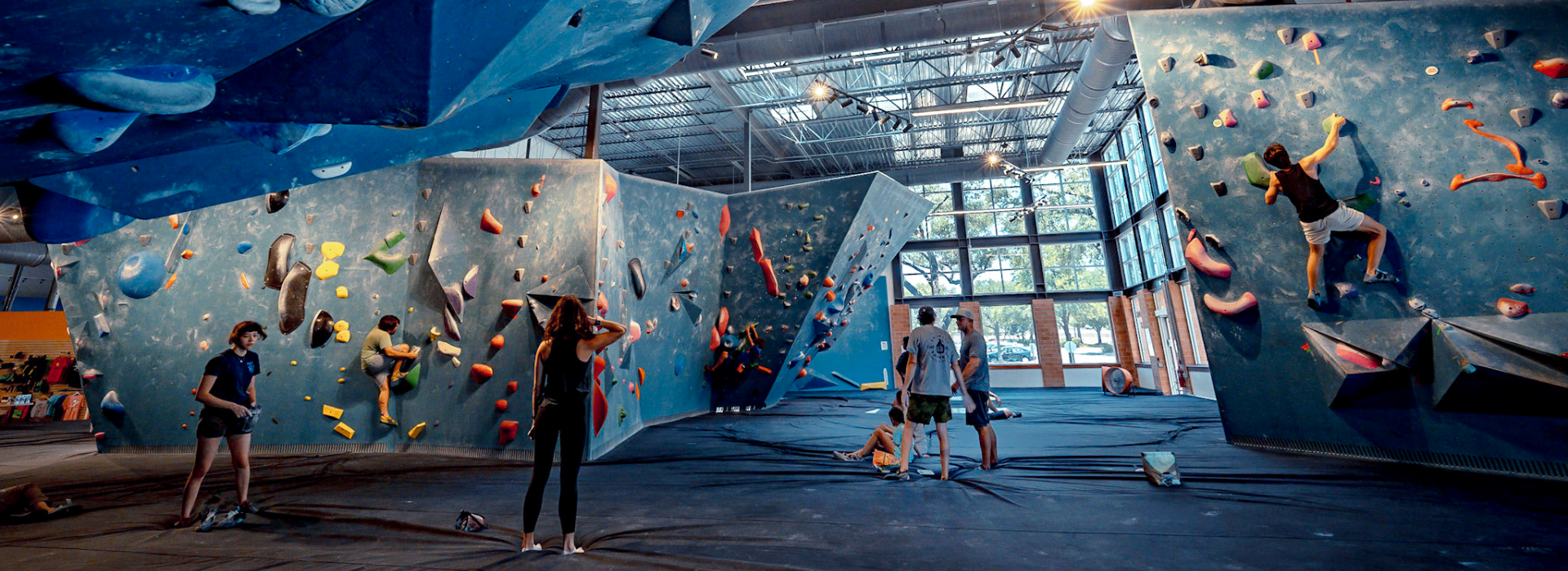 People are indoor rock climbing on bouldering walls with colorful holds at an Austin Climbing Gym. Some climb while others stand, sit, or prepare nearby. Large windows let in natural light, and the gym has high ceilings and padded floors.