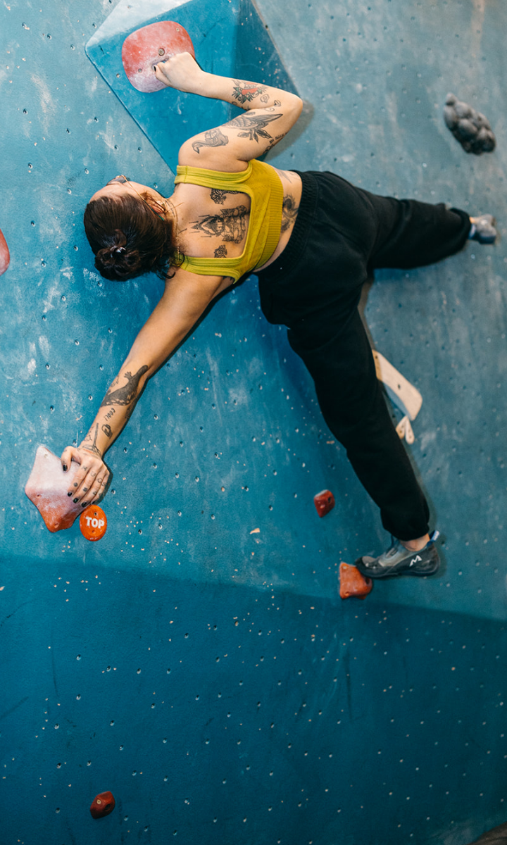 A woman with tattoos wearing a yellow sports bra and black pants showcases her climbing experience as she scales an indoor rock wall, stretching to reach red holds on the blue surface.