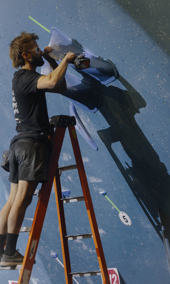 A person stands on a ladder, using a power drill to adjust blue climbing holds on an indoor climbing wall, ensuring the best climbing experience for enthusiasts as route indicators mark the way.