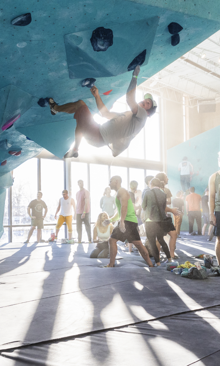 A person climbs an indoor bouldering wall under bright sunlight, while several people watch and gather below on padded mats. The gym is busy, with sunlight streaming through large windows.
