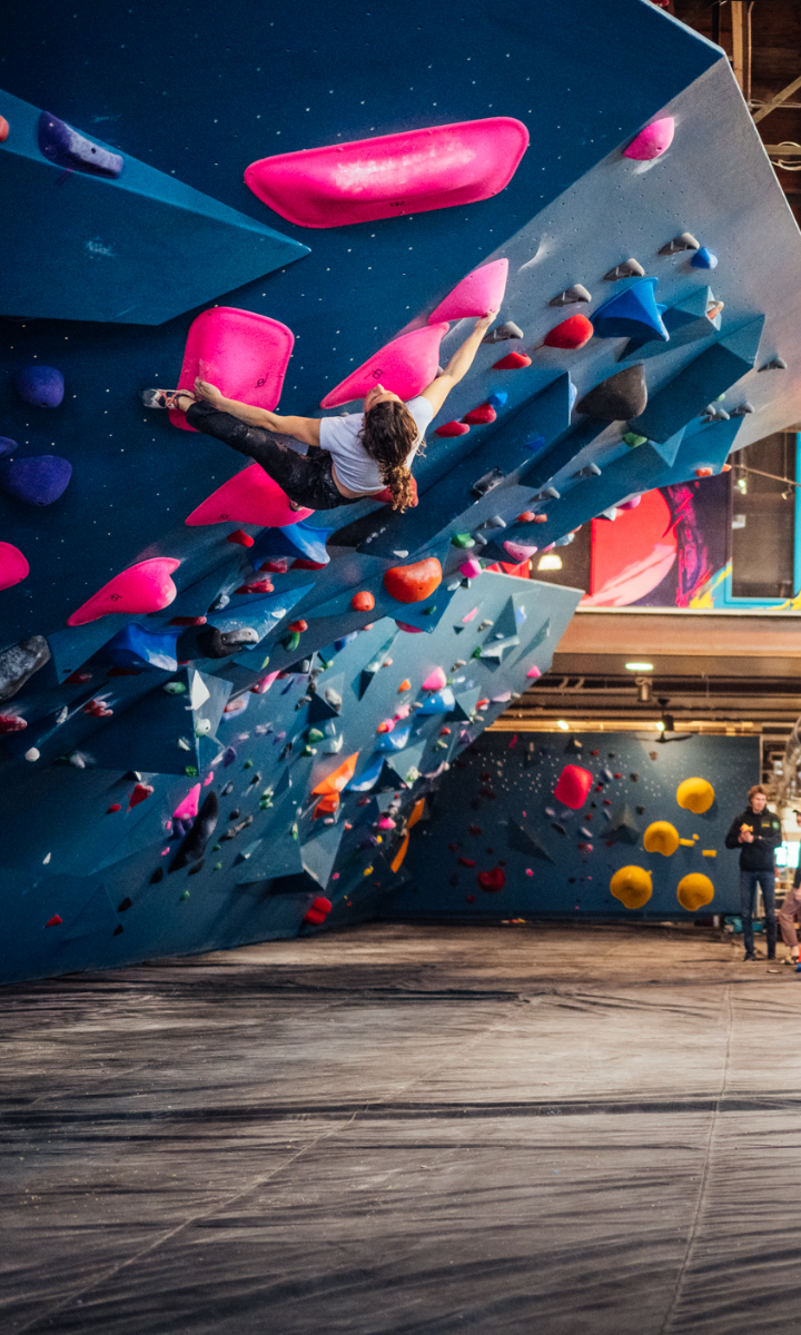 A person with long hair climbs an indoor bouldering wall, gripping large pink handholds on an overhanging section. Other climbers and colorful holds are visible in the background.