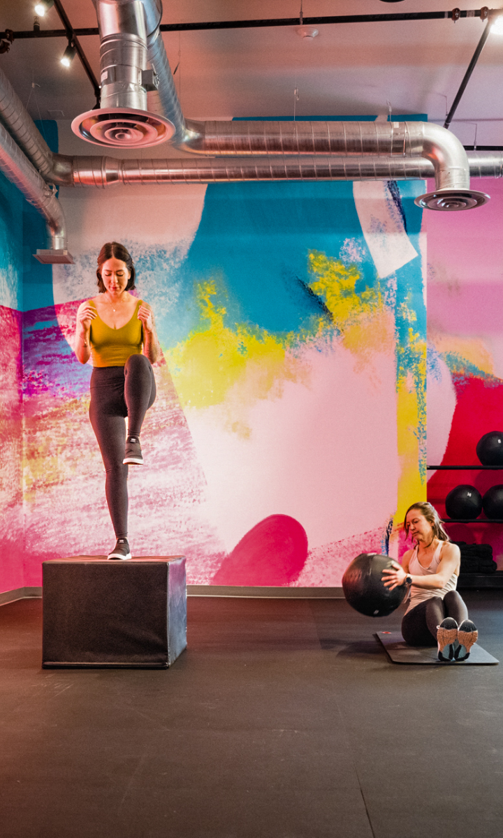 Two women exercising in a colorful gym. One woman steps onto a box while the other sits on the floor holding a medicine ball. Bright, abstract mural and black exercise balls in the background.