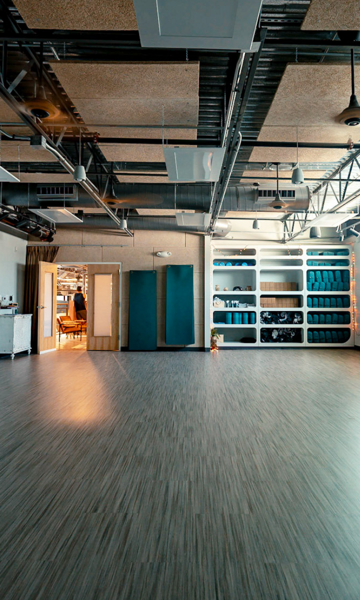 A spacious, modern studio in an Austin Climbing Gym with wooden flooring, shelves holding rolled yoga mats and equipment, and large blue wall pads. Ceiling displays exposed ducts and lighting fixtures. Natural light enters from a glass door.