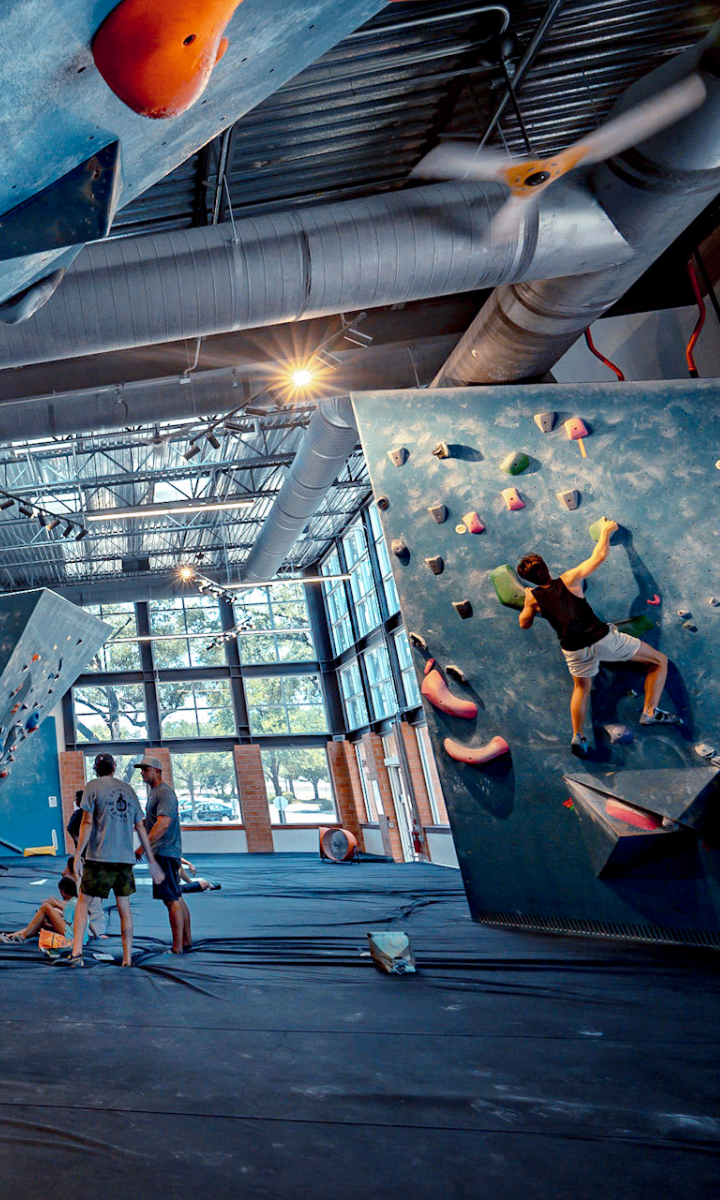 A person climbs a bouldering wall inside a modern Austin climbing gym, while three people stand nearby on padded flooring. Sunlight streams through large windows, and colorful climbing holds are visible on the walls.