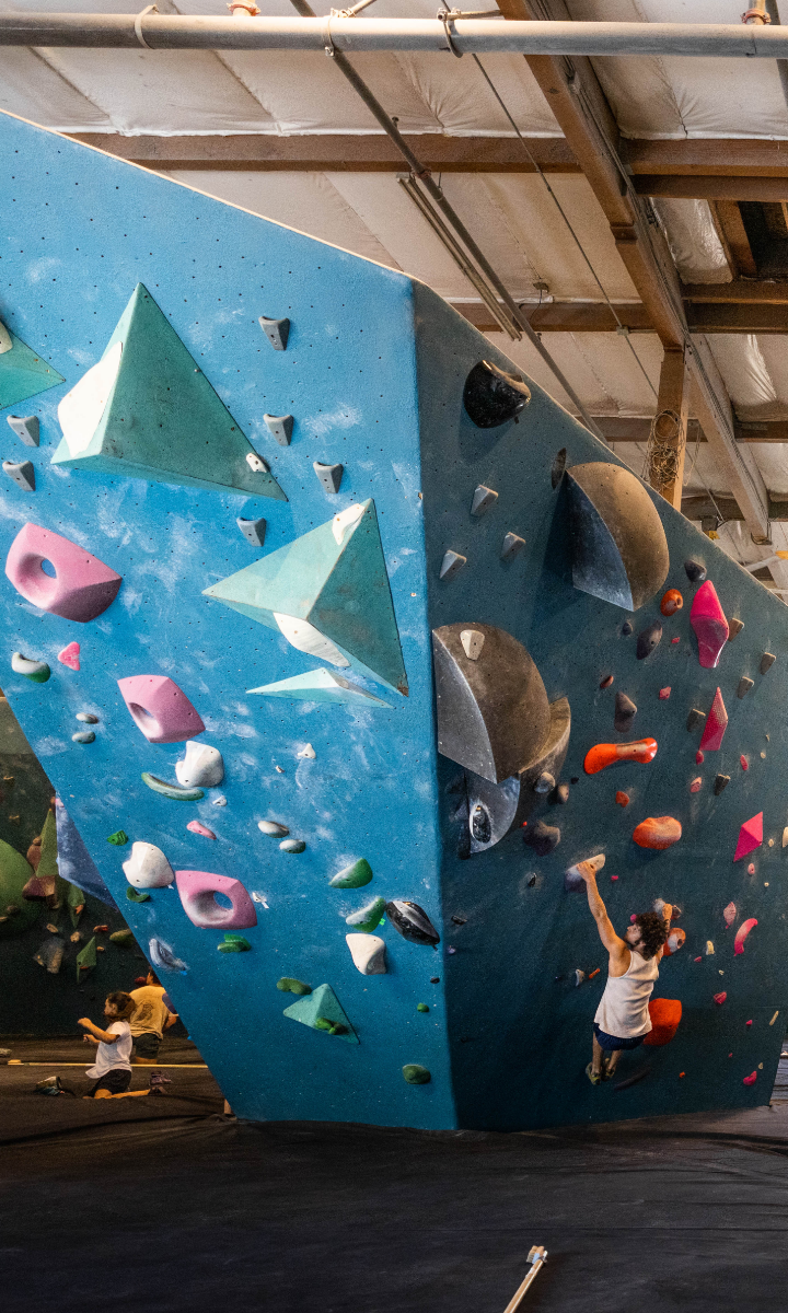 A person climbs an indoor bouldering wall with colorful holds and geometric volumes, reaching up with one arm. The climbing gym has padded floors and exposed ceiling beams.