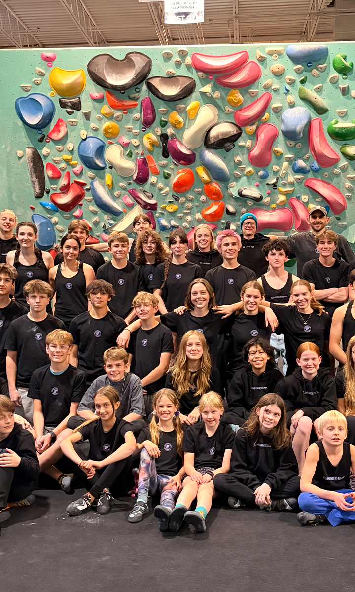 A large group of kids and teens from the Minneapolis Climb Team pose together in front of a colorful indoor climbing wall, all wearing matching black shirts and smiling at the camera.