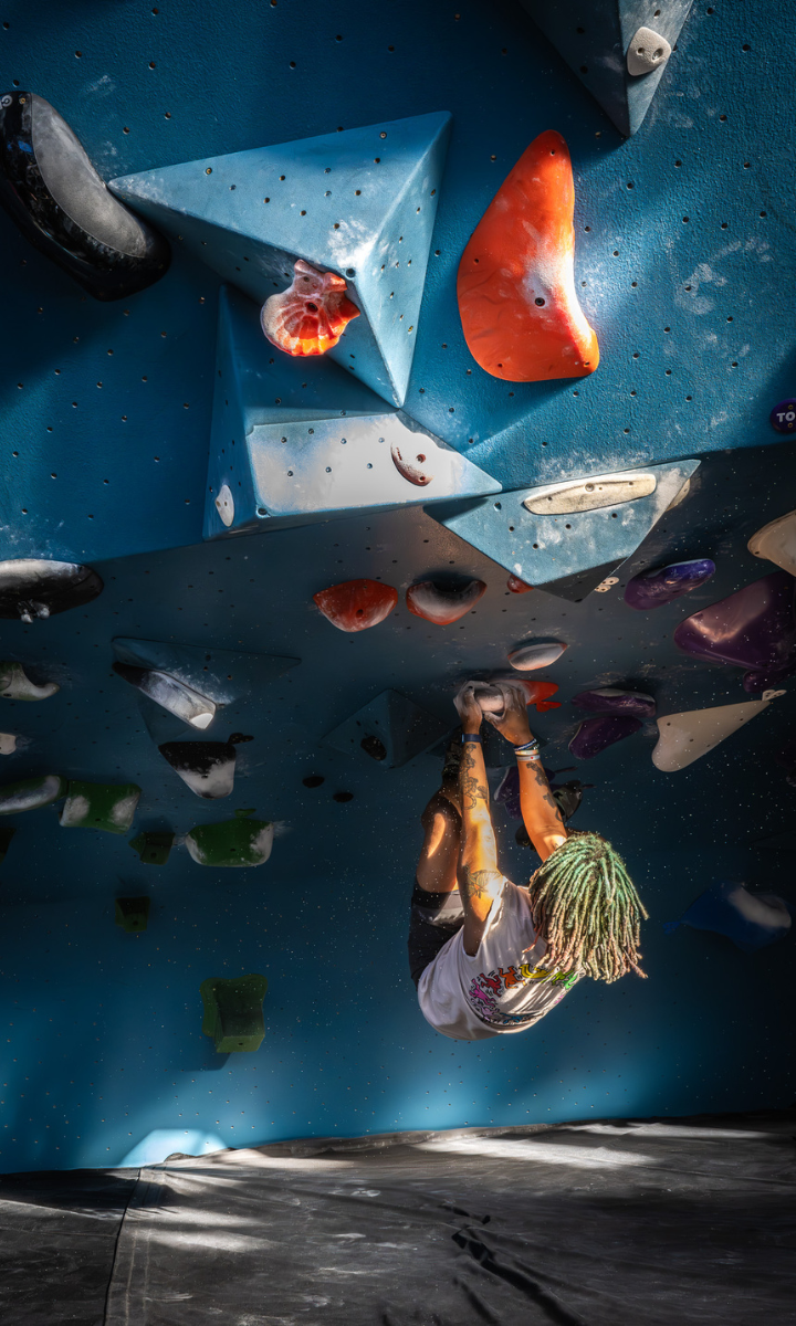 A person with green dreadlocks climbs an indoor bouldering wall, gripping colorful holds on an overhanging blue surface. Bright sunlight highlights their figure at one of todays exciting climbing events in Brooklyn.