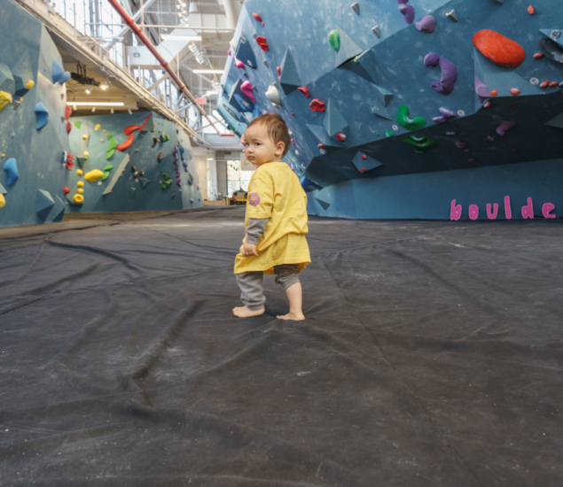 A young child in a yellow shirt and gray pants stands barefoot on a black mat inside an indoor climbing gym with colorful climbing holds, showcasing the vibrant atmosphere of the Youth Program.