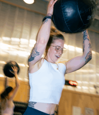A woman with glasses and tattoos lifts a medicine ball overhead during a workout in a gym. Another person in the background is performing a similar exercise. The setting is bright with blurred motion, showing intensity.