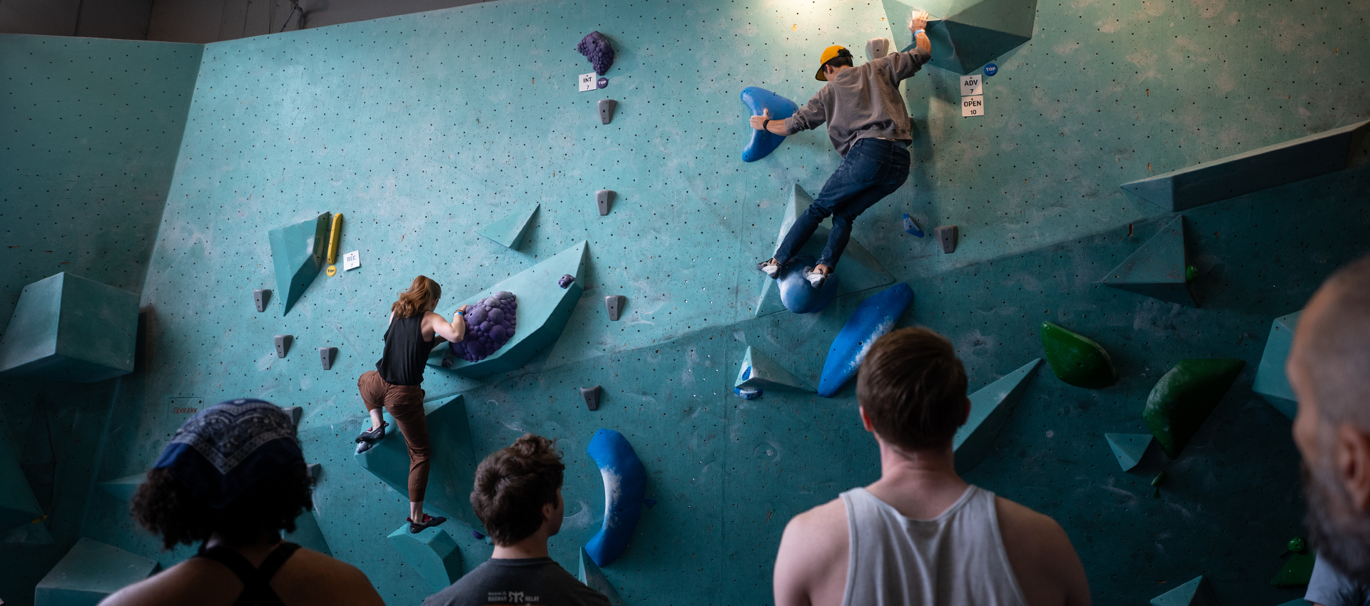 Several people watch as two climbers ascend a blue indoor bouldering wall with various colored holds and volumes. The climbers are focused; the observers stand in the foreground with their backs to the camera.