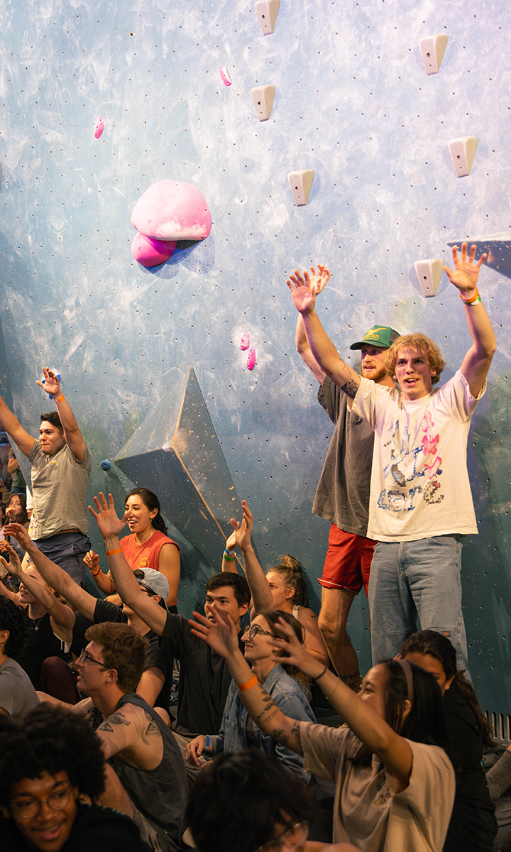 A group of people sits and stands in front of an indoor climbing wall, cheering with raised arms and smiling faces. The blue wall, part of the Climb for Community event, features pink climbing holds and geometric shapes.