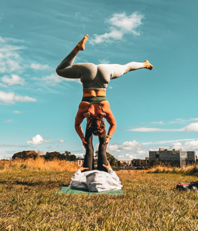 Two people practice acro yoga outdoors on a grassy field. One person lies on their back with legs up, supporting the other, who balances upside down on their hands. Buildings and trees are visible in the background under a blue sky.