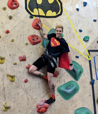 A person wearing a Superman cape and T-shirt poses while rock climbing indoors. They are smiling, one leg up on a green hold, with a Batman sign above them and colorful climbing holds on the wall.
