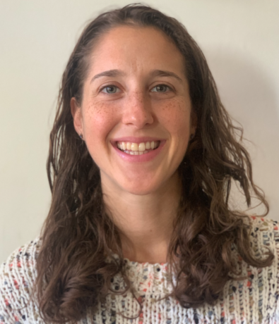 A woman with long brown hair and a light complexion smiles at the camera. She is wearing a white knit sweater with colorful specks, standing in front of a plain light background, looking ready for her next climbing adventure.