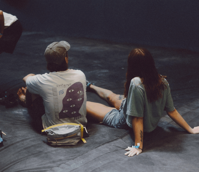 Two people sit on a dark surface, facing away from the camera. One wears a cap and a T-shirt with a graphic of multiple eyes; the other has long hair and a T-shirt. A small bag with a BP logo rests on the ground beside them.