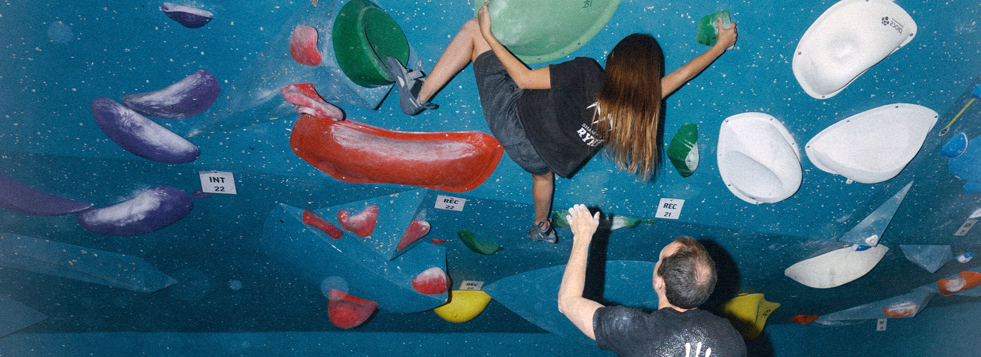 A woman climbs an indoor bouldering wall with colorful holds while a man stands below, watching and spotting her. Both wear black shirts in a brightly lit gym offering Personal Climbing Coaching Austin.