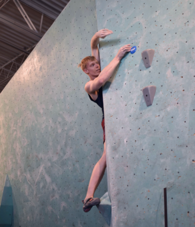 A person with light hair climbs an indoor bouldering wall in Minneapolis, reaching high with one arm while balancing on small footholds—an ideal challenge for those seeking private coaching. The wall is light blue with scattered handholds and a speckled texture.
